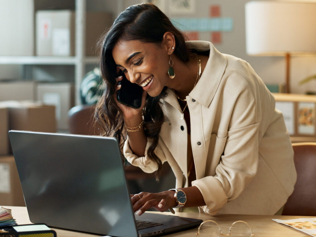 Home Female business owner on her mobile phone to HR consutant whilst working on laptop. In the background there are customer packages ready to be sent.